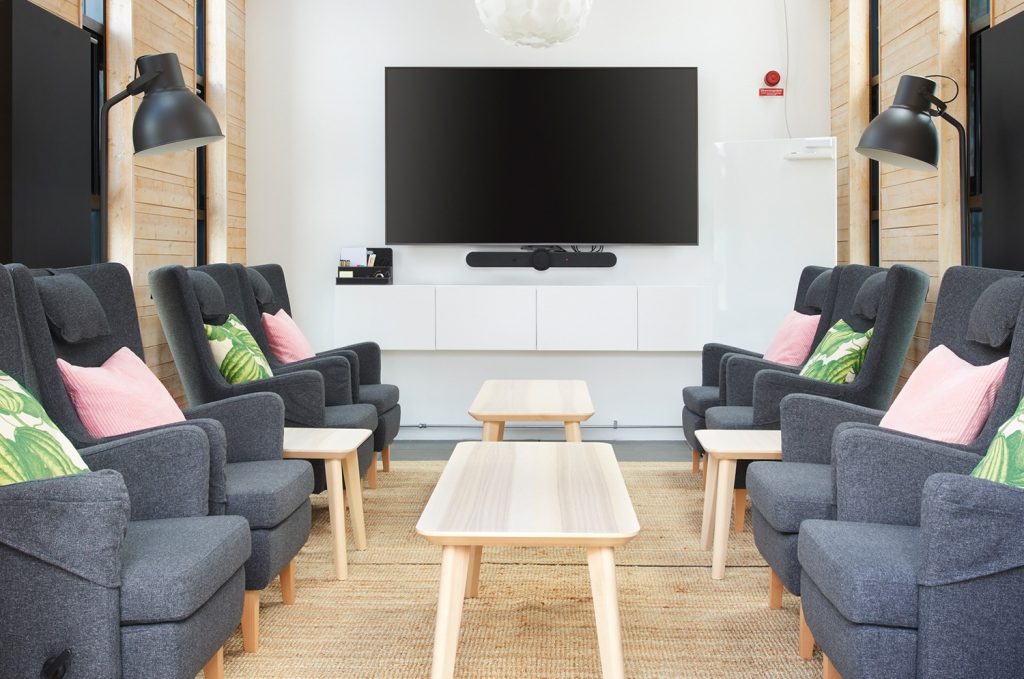 Eight armchairs on a big carpet with coffee tables in a high ceiling meeting room, with plenty of windows and a big tv screen.