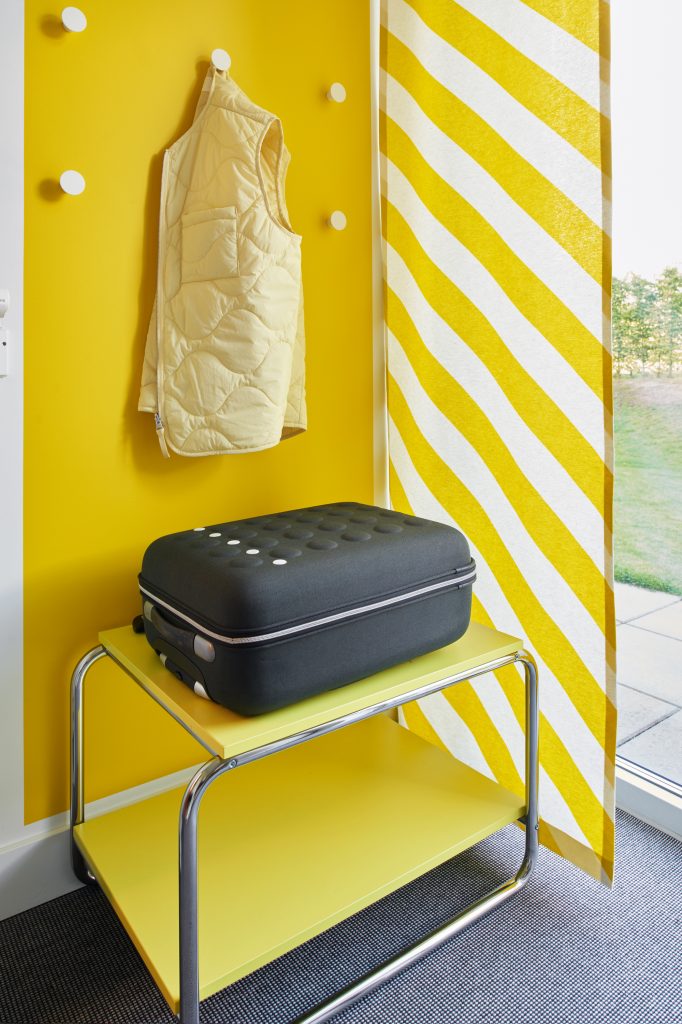 Ett gult nattduksbord med en rullväska ovanpå och krokar för kläder på väggen bakom.