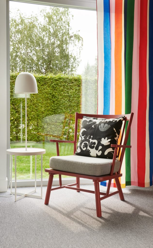En röd fåtölj och en golvlampa framför ett stort fönster med färgglada gardiner.