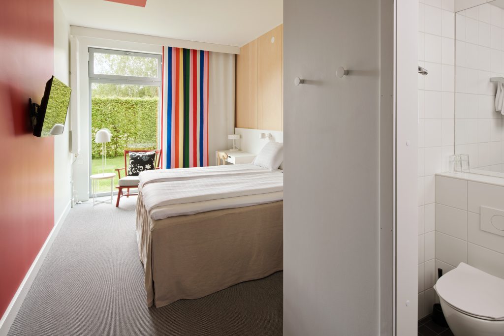 En överblick av ett dubbelrum, med en TV, dubbelsäng, fåtölj och öppen dörr till badrummet.