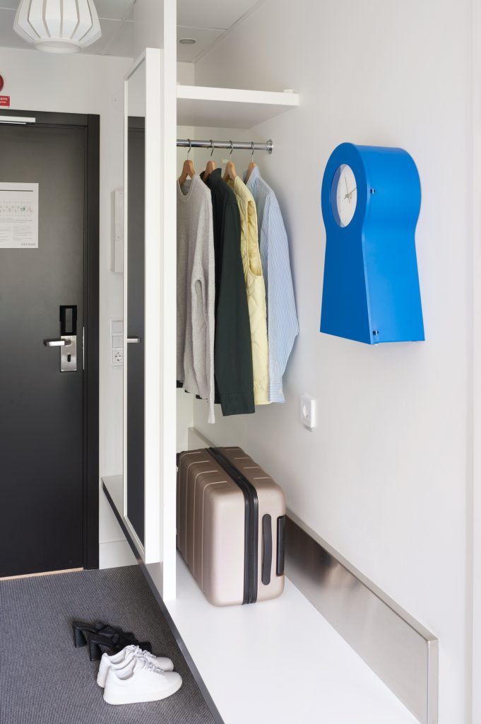 Förvaring för skor, rullväska och kläder på galgar med en blå klocka monterad på väggen bredvid.